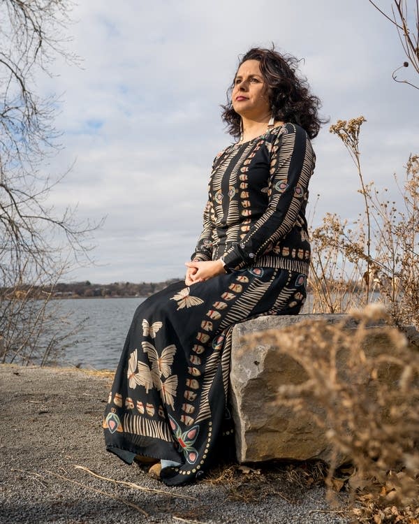 A woman in a patterned dress sits on a rock in front of a lake.