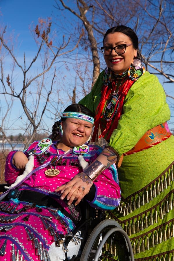 woman stands with daughter in wheelchair wearing traditional regalia